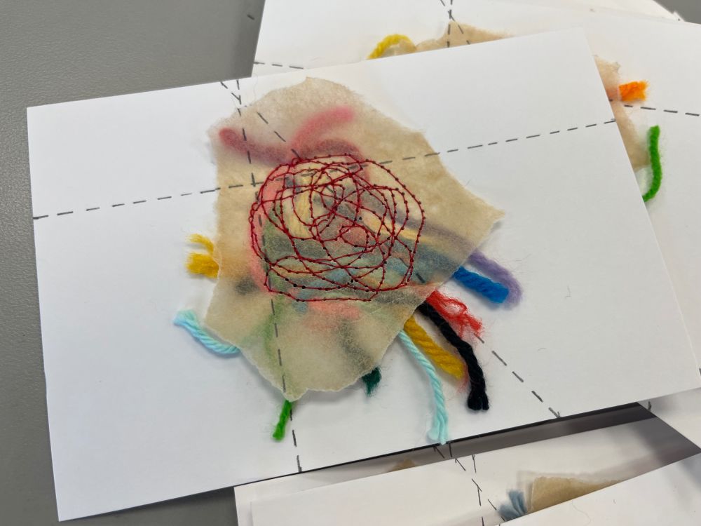 Photograph of a white postcard with colorful yarn scraps placed in the center, covered with a small torn piece od brown parcahment paper. There is freeform stiching in red thread that goes through the parchment and yarn attaching it to the white postcard. Three dashed lines, in pencil, have been drawn across the postcard, parchament paper, and stitching.