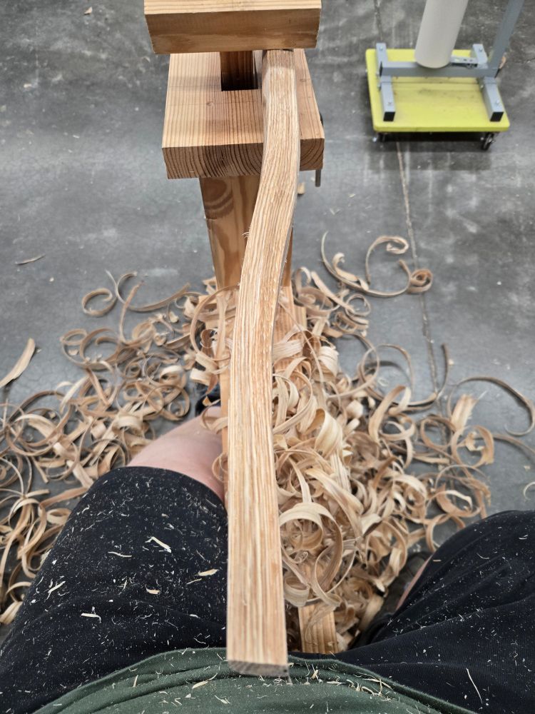 Seated at a shaving horse and surrounded by piles of curly wood shavings - a naturally curved piece of white oak is clamped into the horse and has been shaved smooth with a draw knife.