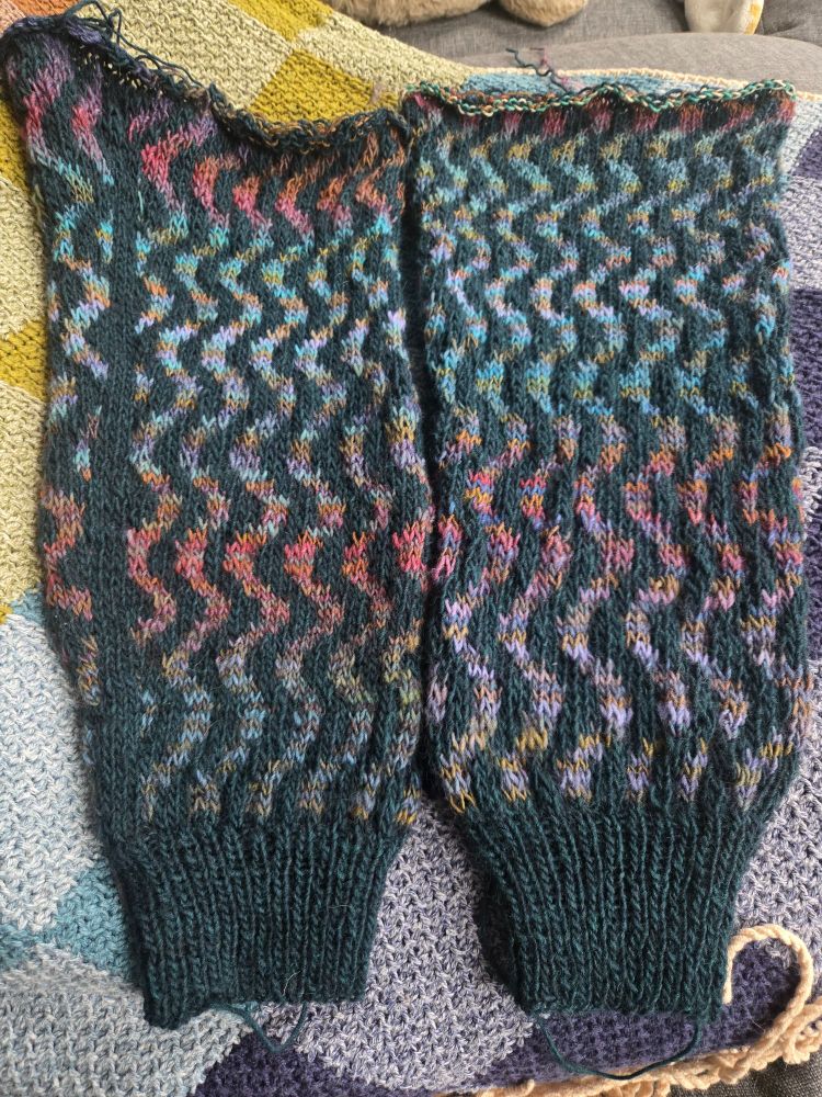 Two hand knitted sweater sleeves, side by side on a blanket. They feature a squiggle colorwork pattern in deep teal and a variegated yarn in shades of red, purple, and green.