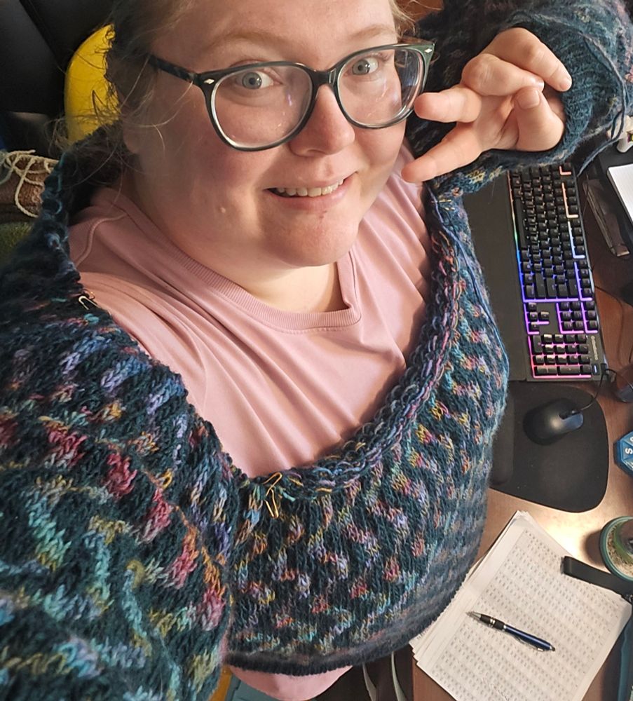 Trying on a partially finished sweater in my office. Pattern is Partner Pullover by Lydia Morrow in size 9. Yarn used is Hobbii Winter Glow in col 21 (a subtly heathered multicolor in muted shades of red, yellow and teal) and Hobbii Winter Glow Solid in Petrol (a deep teal).