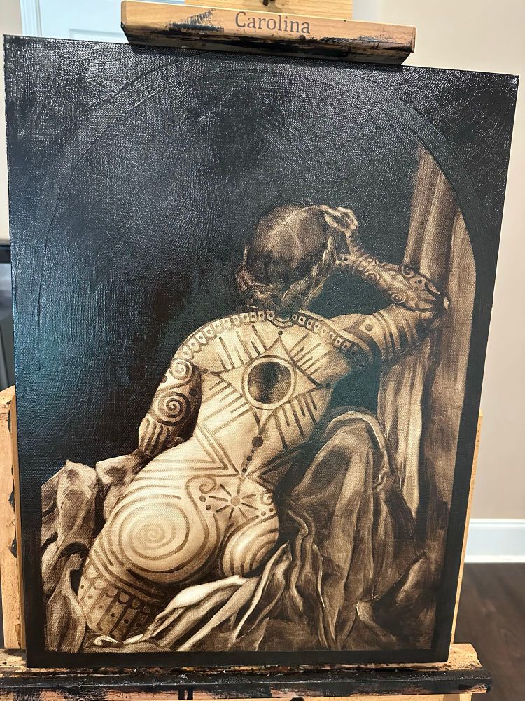 Image of an oil painting resting on an easel of a nude woman’s back. She is reclining on fabric with her arm raised. She has bold ornamental tattoos over her entire back, arms, and legs. Image is reminiscent of vintage photographs of tattooed women at freak shows.