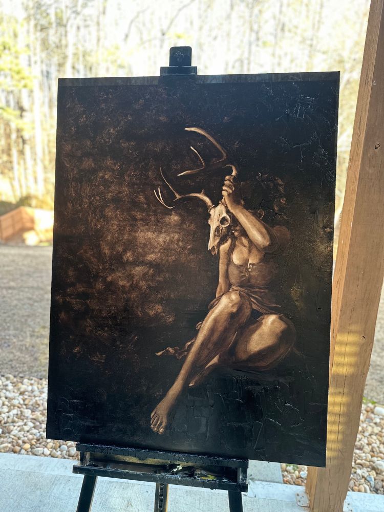 Image of a large oil painting on an easel outdoors of a woman holding a deer skull in front of her face. The woman is painted in a realistic style and the background is dark with abstract textures over it. There’s thick chunks of paint in the corners where the dark sepia tone is mostly black