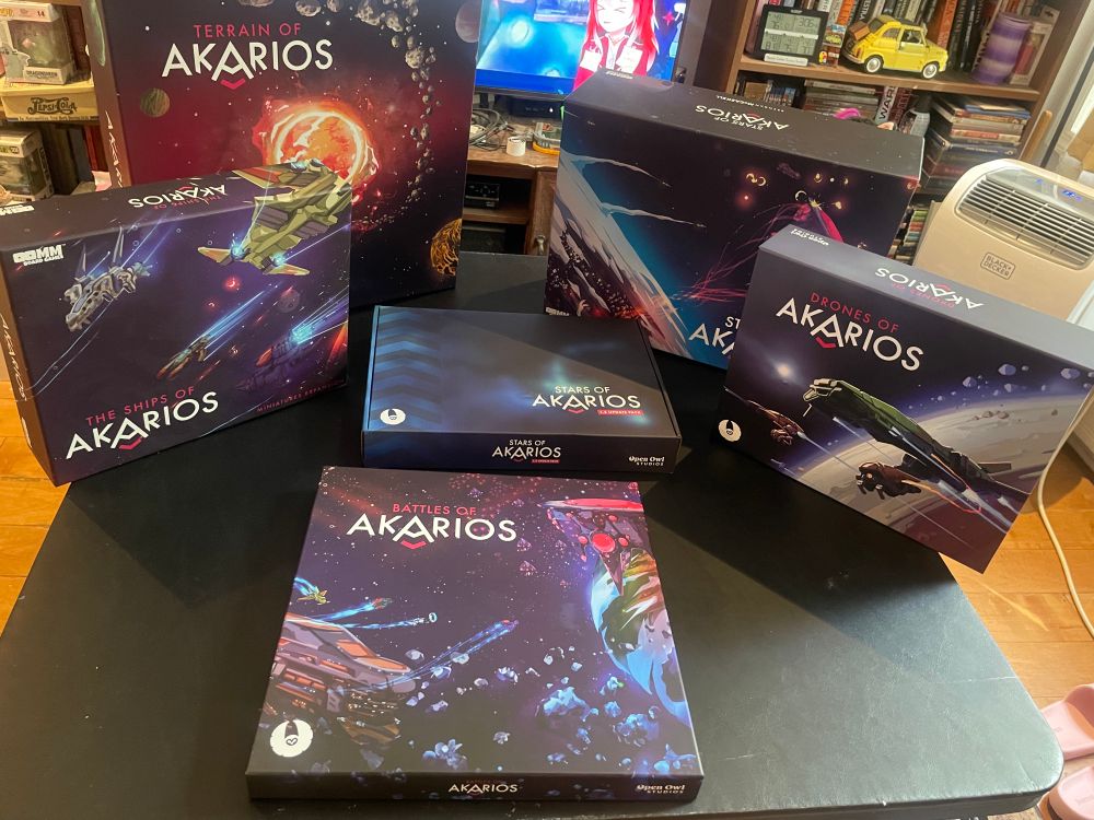 6 boxes on a card table. The boxes contain the various components of Stars of Akarios. The base game and the 1.5 update boxes. 