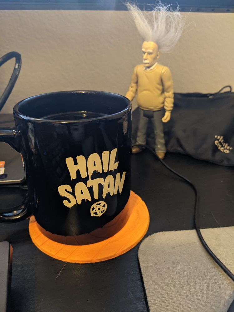 A picture of a coffee cup with hail Satan written on it and an Einstein action figure in the background.