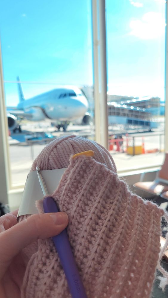 My hand holding up my work in progress crochet project. The yarn is pink and the crochet hook is purple. In the background is a window and an airplane sitting on a tarmac. 