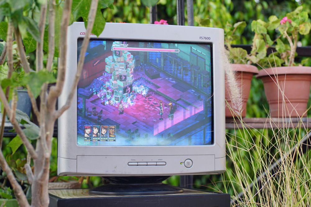 Demonschool, specifically a boss fight in a purple environment featuring a stack of televisions, is displayed on a white compaq 4:3 CRT on a table in a roof garden surrounded by plants.