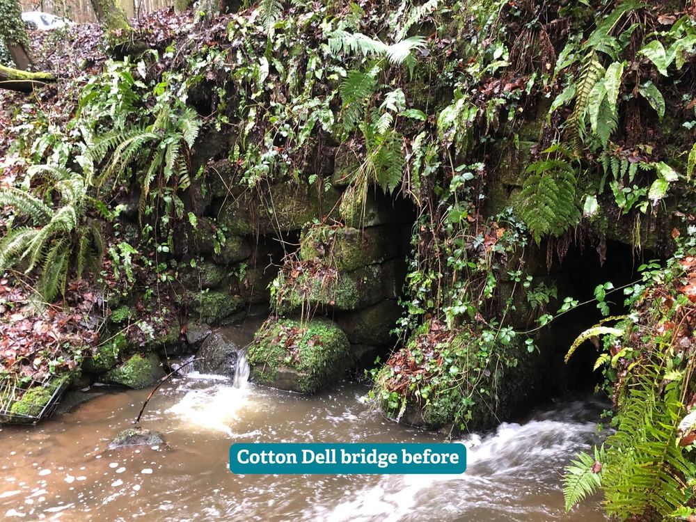 An old crumbling stone bridge covered in moss with a stream running under it. The stones are falling down and it looks unstable.