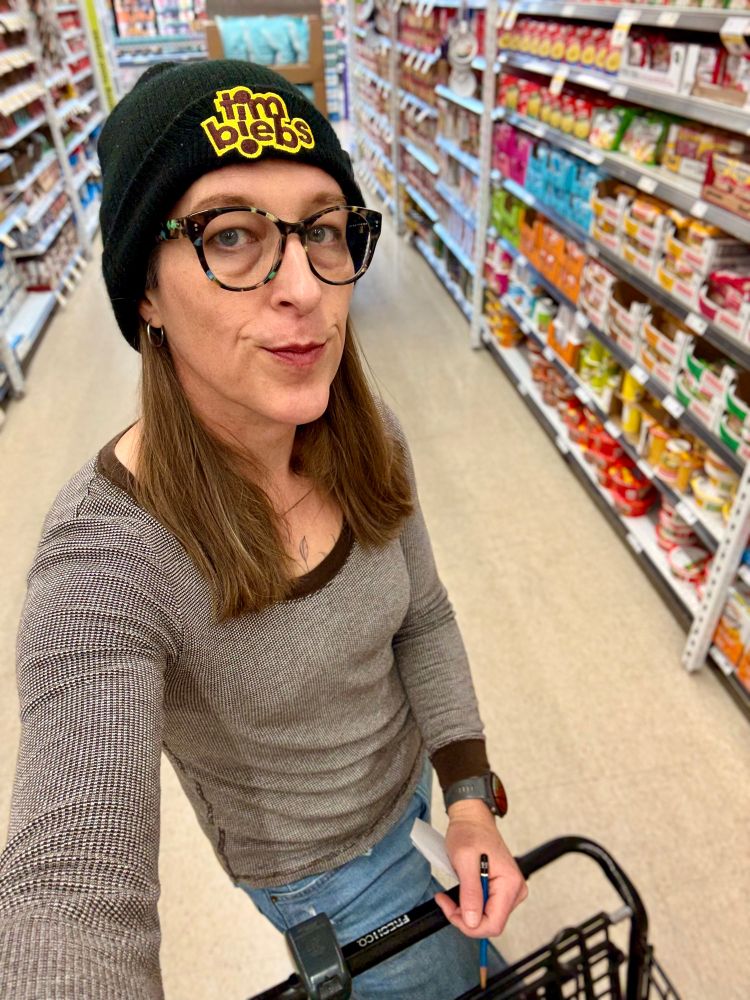 Standing in a grocery store aisle wearing jeans, long sleeve brown top and a black toque