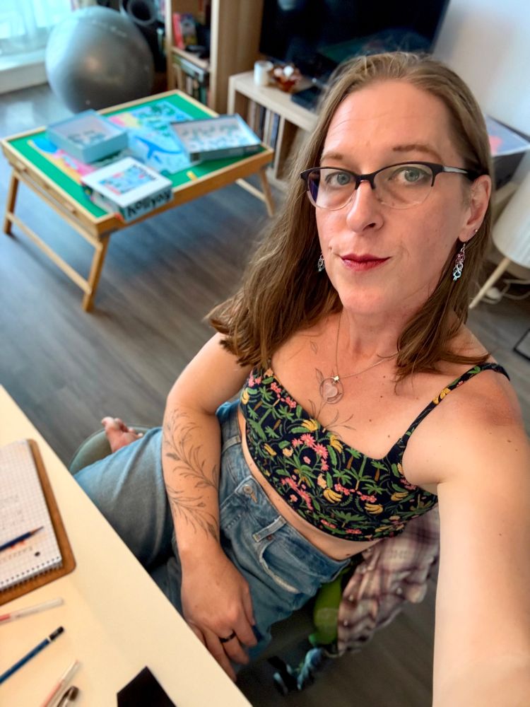 Pic of me sitting at my desk. I’m wearing ripped jeans with one leg under the other on my chair. I’m also wearing a brami with a tropical pattern on it. My puzzle table is in the background