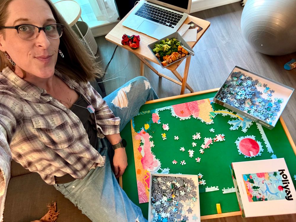 I’m holding my camera up to catch the scene of me sitting on the couch with a puzzle table in front of me between my legs. The puzzle is only partially completed. There a tv tray beside me with a laptop and a bowl of food. I’m wearing ripped jeans, a dark grey tank top and a white and purple checked shirt