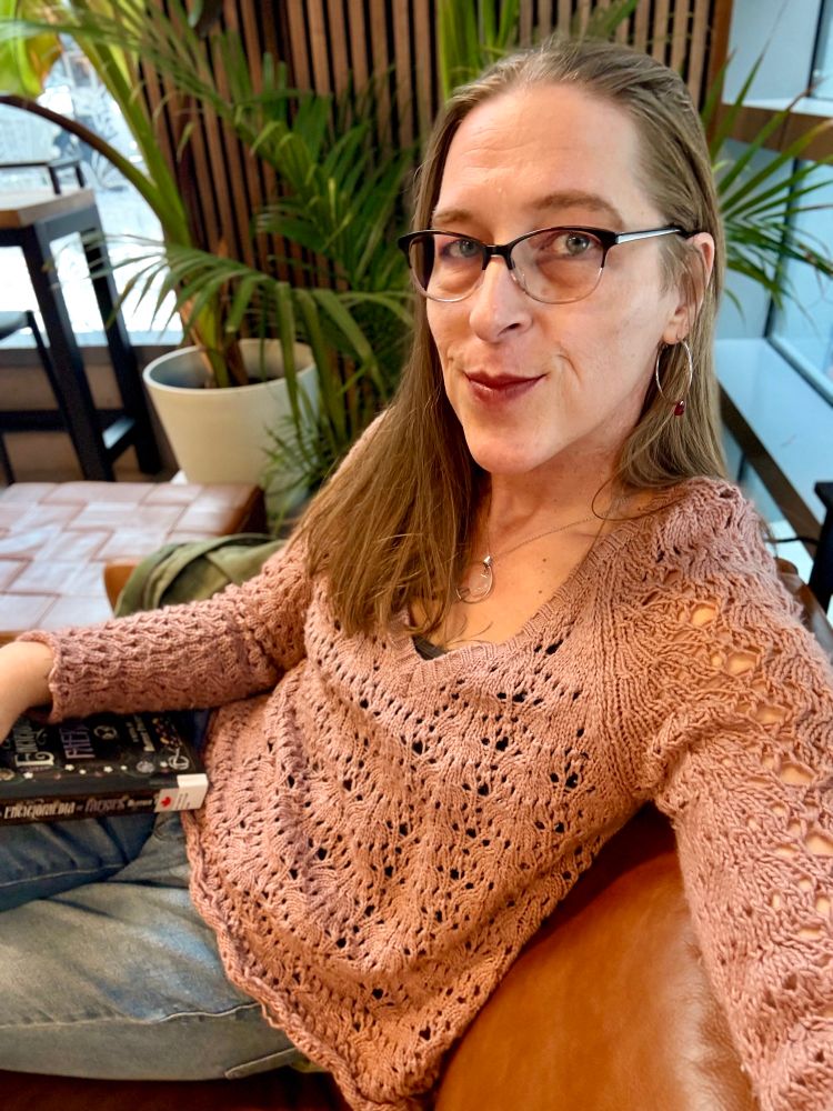 Sitting on a brown leather couch at a cafe with plants behind me. I’m wearing jeans and a pink open knit sweater