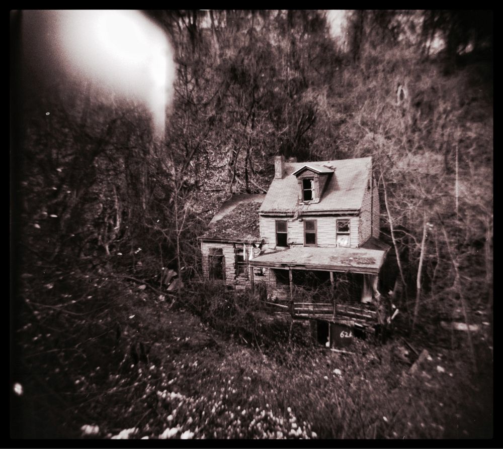 A dilapidated house that’s falling apart, nestled into an overgrown hillside. Image is black and white and sort of dreamy looking thanks to the use of a Holga. An imperfect camera that produces interesting results.