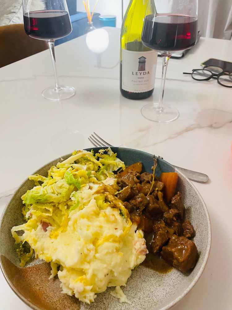 A plate of beef and Guinness stew with colcannon and extra cabbage. A glass of red wine in the background. 