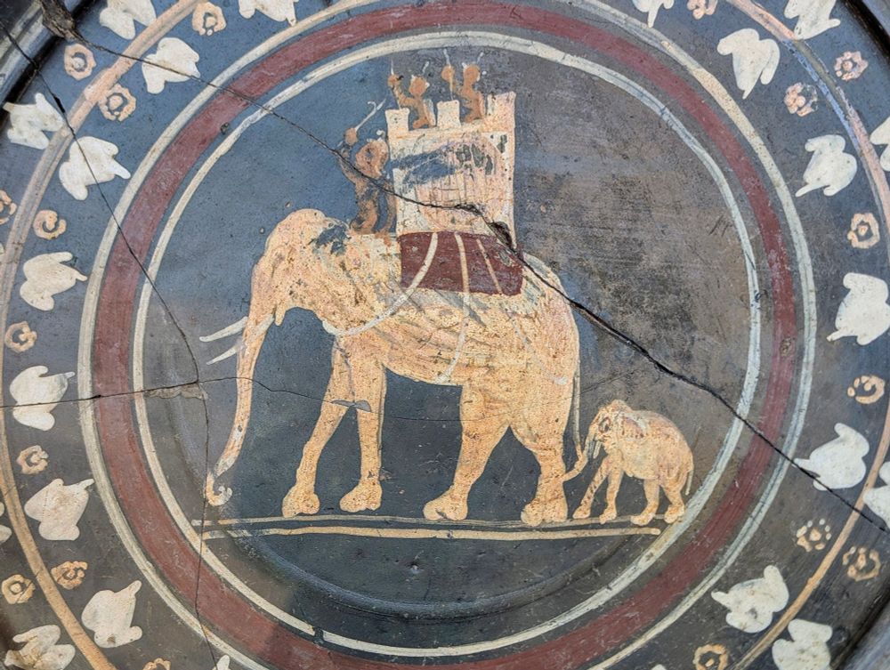 A figuratively decorated plate with black gloss, showing a war elephant with a baby elephant following behind, perhaps in a parade.