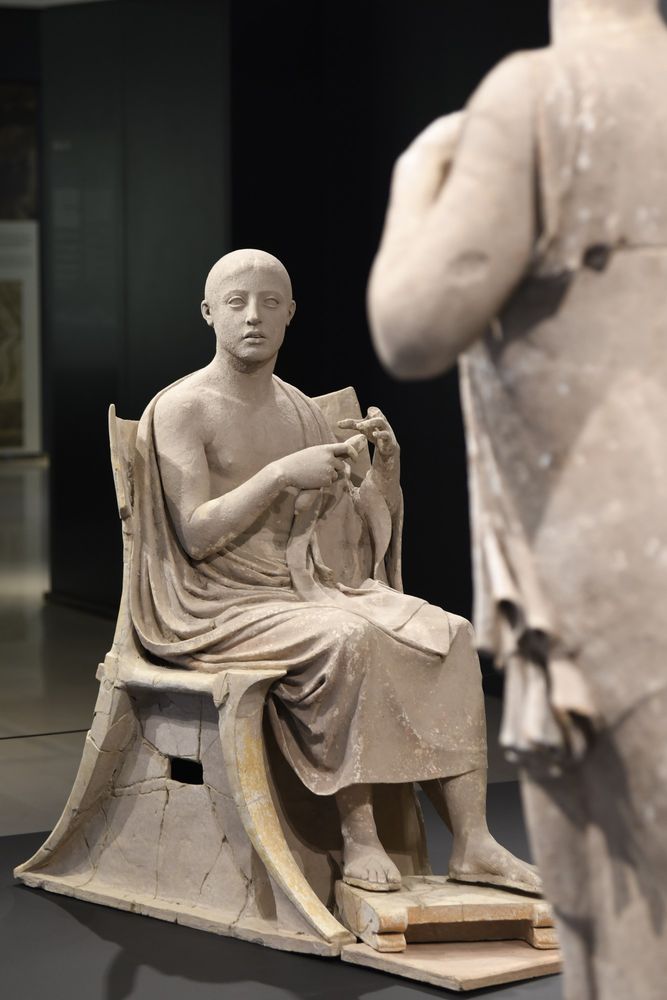 Three terracotta statues of Orpheus, sitting on a stool and playing his instrument, and two sirens, half human and half bird, standing in front of him.