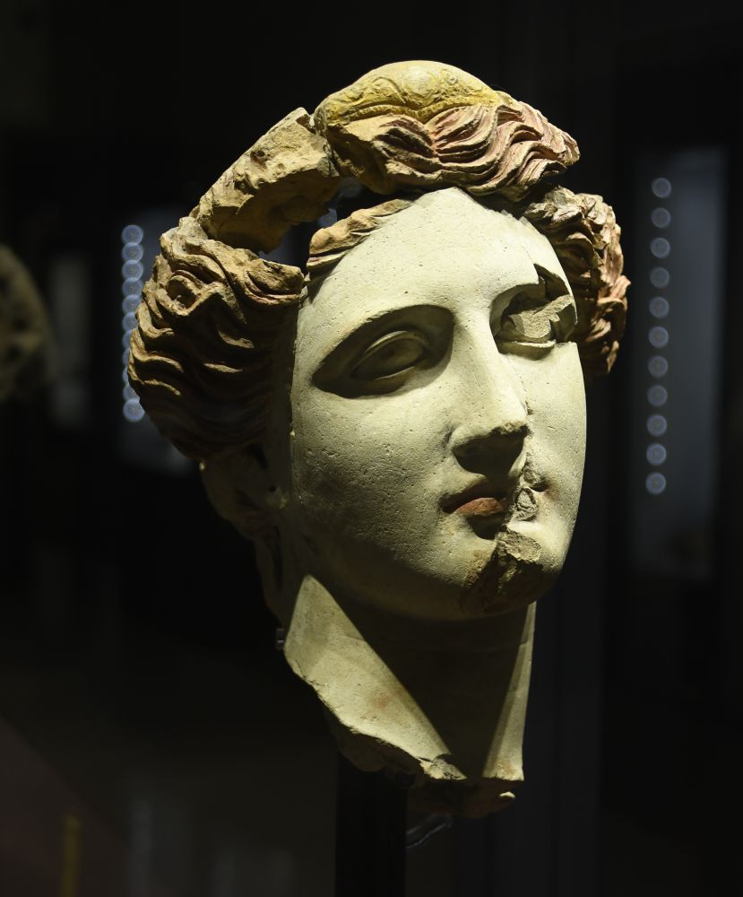 A female head with a neutral expression, red lips, reddish-brown hair and a yellow diadem imitating gold.