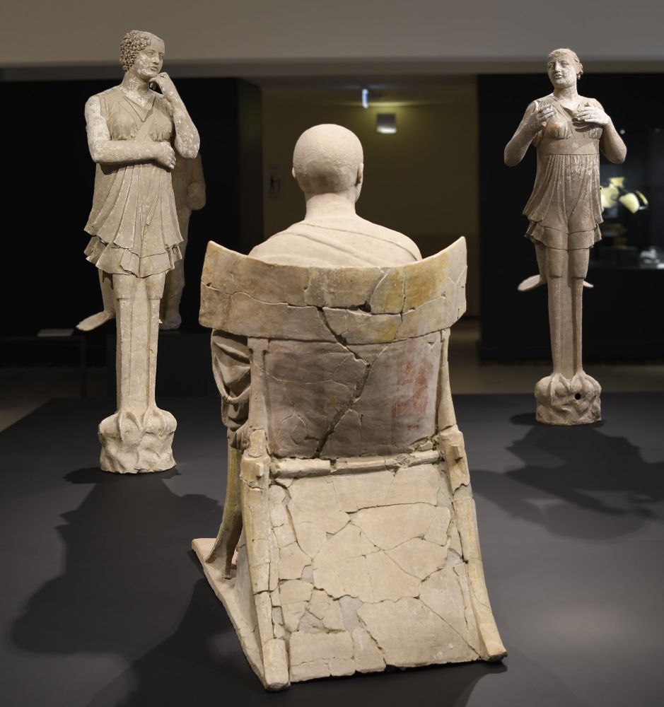 Three terracotta statues of Orpheus, sitting on a stool and playing his instrument, and two sirens, half human and half bird, standing in front of him.