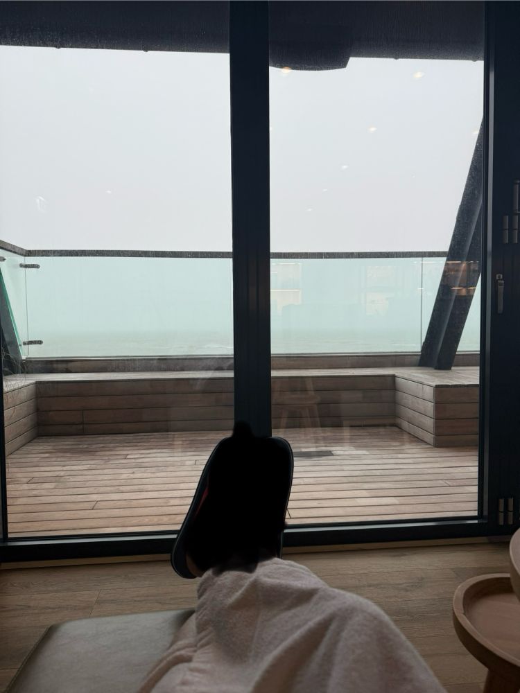 Verpixelte Füße, Holzboden, Fensferfront, im Hintergrund Wasser, Wellen und grauer Himmel.