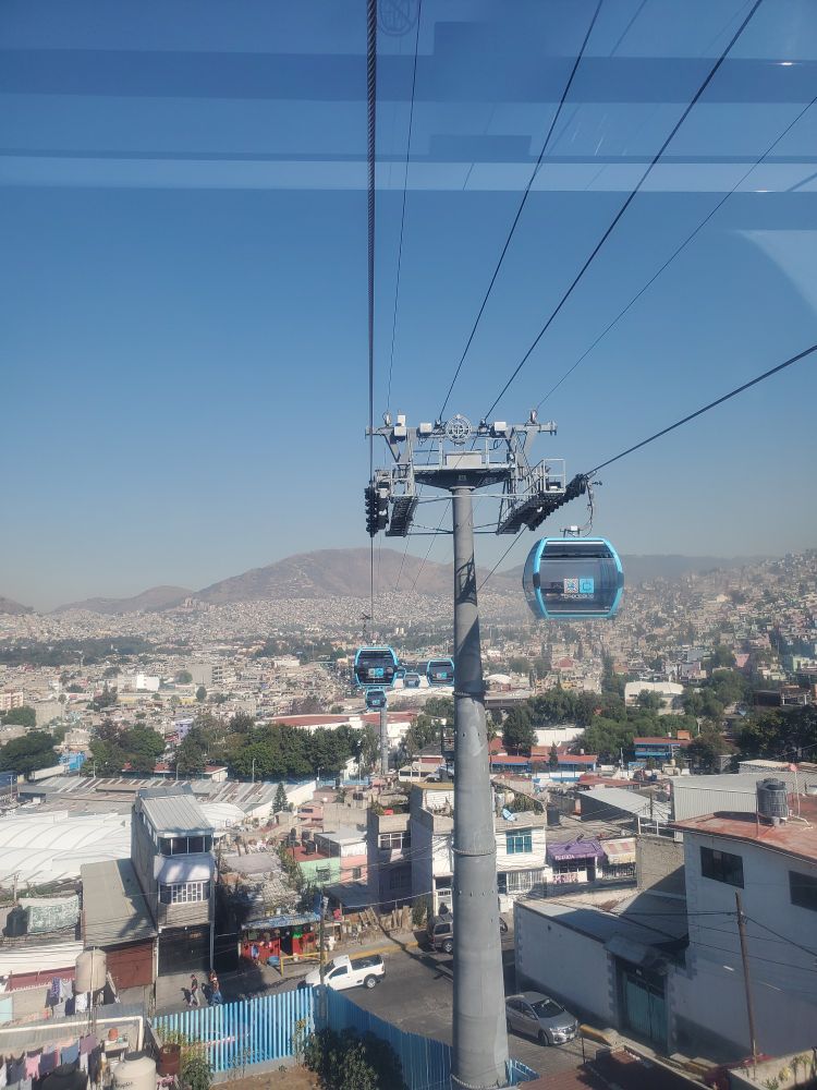 Blue gondolas floating above a busy metropolitan of cement houses. Rounded mountains form along the horizon. 