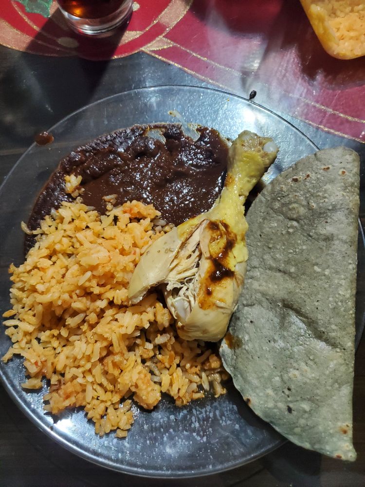 A glass plate with yellow Mexican rice, chicken drumstick, dark brown mole, and a blue corn tortilla. 