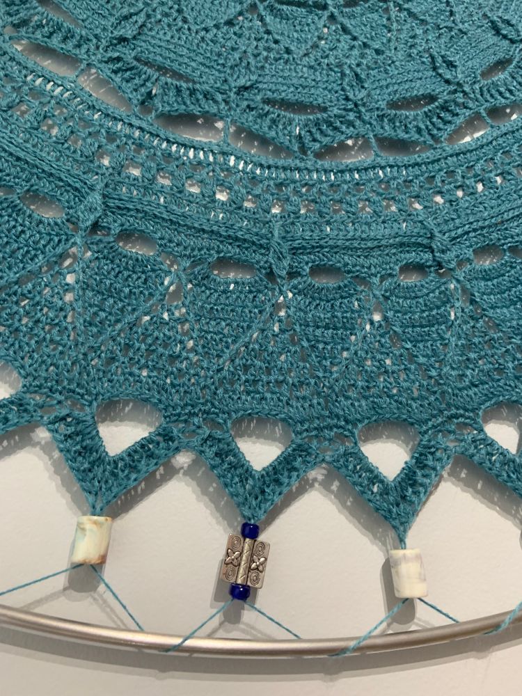 Close up of the stitch detail of a crochet wall hanging in blue/green 4 ply yarn (mystery yarn purchased at auction years ago) attached to a steel ring with ceramic and metal beads hangs on a grey wall. Pattern is Mandala Flores by Desi Ven  
https://ravel.me/mandala-flores

I used a 2.5 mm crochet hook, the beads were purchased from Shipwrecked Bead Shop in Courtenay BC. The steel hoop is 1/4 inch thick x 42 inch circumference from Unfettered on Vancouver Island https://unfetteredsupply.com/