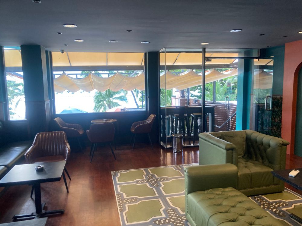 An arrangement of brown and green chairs and tables with ashtrays for enjoying cannabis. The ocean out the window can’t really be made out due to the light.