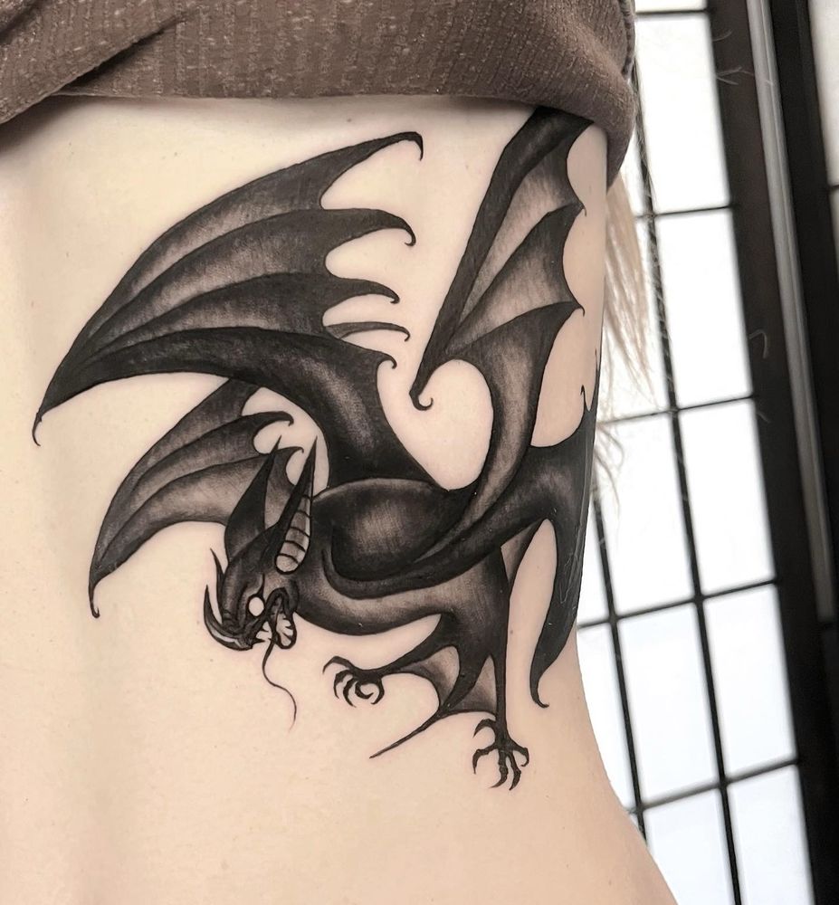 A black and grey tattoo of a 4 winged bat with a serpentine tongue. The tattoo is on the side of the torso on the ribcage, wrapping towards the front of the stomach
