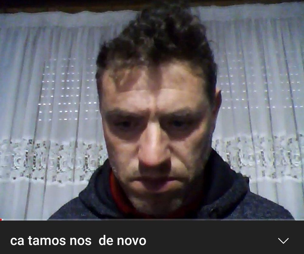 print de um frame de um vídeo no qual está enquadrado um homem branco maduro com vincos na testa e no rosto e com cabelo curto, escuro e um tanto despenteado. ele traja um moletom escuro e atrás dele há uma cortina branca. sombras pairam sobre seus olhos e boca, dando-lhe uma aparência sorumbática. o título do vídeo é "ca tamos nos  de novo" (sic)