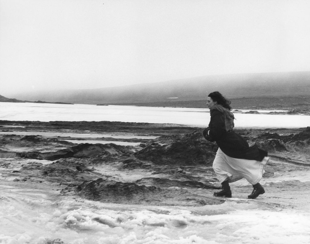 A white woman in a long white dress, black overcoat and black boots is walking across a rocky, snowy landscape, looking very cold and windswept and dramatic. It's a still from Sally Potter's film The Gold Diggers.