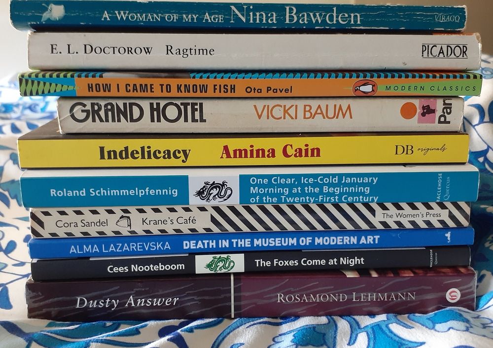 A pile of book spines. From top: A Woman of My Age by Nina Bawden, Ragtime by EL Doctorow, How I Came to Know Fish by Ota Pavel, Grand Hotel by Vicki Baum, Indelicacy by Amina Cain, One Clear, Ice-Cold January Morning at the Beginning of the Twenty-First Century by Roland Schimmelpfennig, Krane's Cafe by Cora Sandel, Death in the Museum of Modern Art by Alma Lazarevska, The Foxes Come at Night by Cees Nooteboom and Dusty Answer by Rosamond Lehmann.