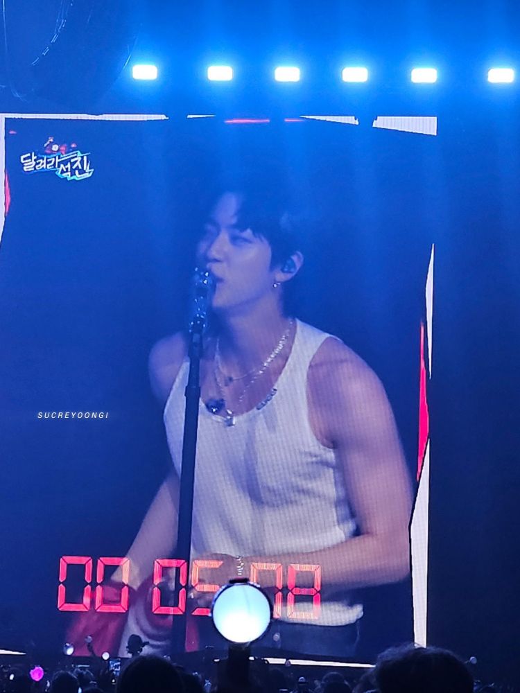seokjin on the jumpotron, he's talking into the mic. he's in a white tank and silver jewelry. he has a red jacket on his hand