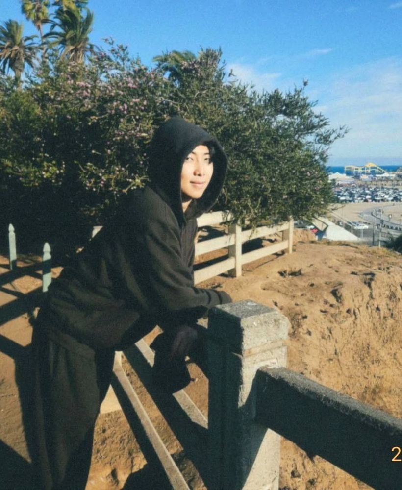 a film photo of namjoon in black hooded sweatshirt and sweat pants leaning against a fence overlooking the beach