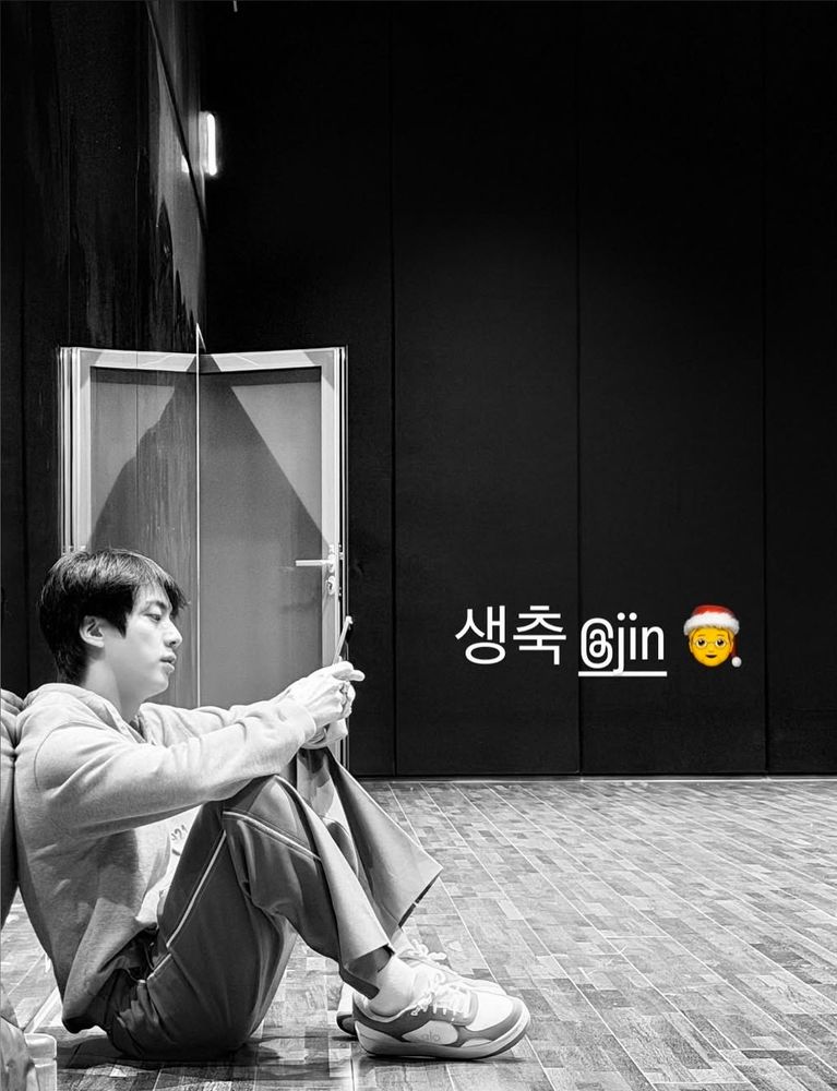 NJ IG story. black and white photo of SJ in comfy clothes sitting on the floor of the practice room and looking at his phone with his back against the mirror