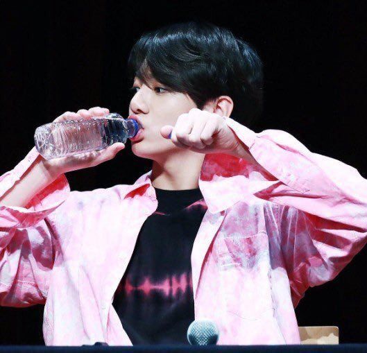 black hair jk in pink tie dye dress shirt with a black tee with a pink the dye? graphic at the chest. he’s holding his left hand up in a fist and the other hand holding a water bottle