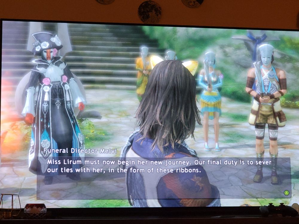 We see the main character of Lost Odyssey, Kaim, from behind. He is wearing yellow cat ears. In front of us to our right are four people in jrpg beach wear in yellows, blues, greens, and browns, wearing strange emotionless white stone masks and holding unlit torches. To the left is a funeral director wearing a flamboyant black, white, and blue costume with a bunch of pink and white roses all over it. Their hands and a stripe down their white carnival face mask are orange. A subtitle for Funeral Director Meivi says: "Miss Lirum must now begin her new journey. Our final duty is to sever our ties with her, in the form of these ribbons."