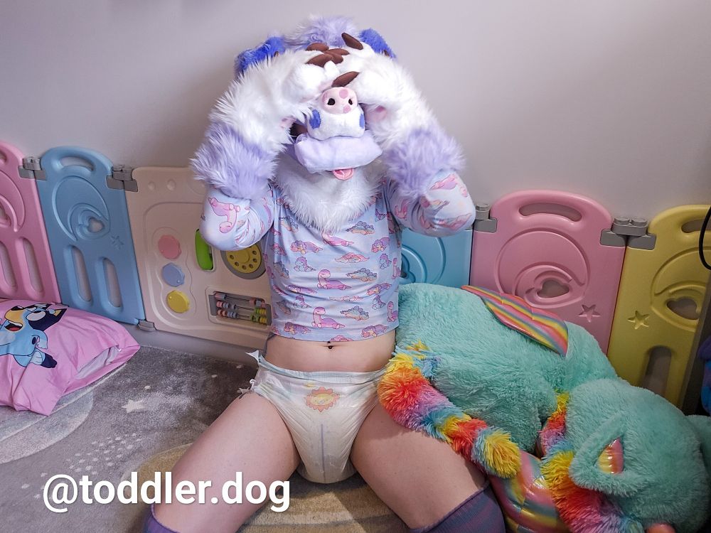 Partial fursuiter border collie dog in a diaper snoofing a pissed in pull up. They are sitting on top of a space themed rug in front of a large dragon plushie and a plastic playpen. 