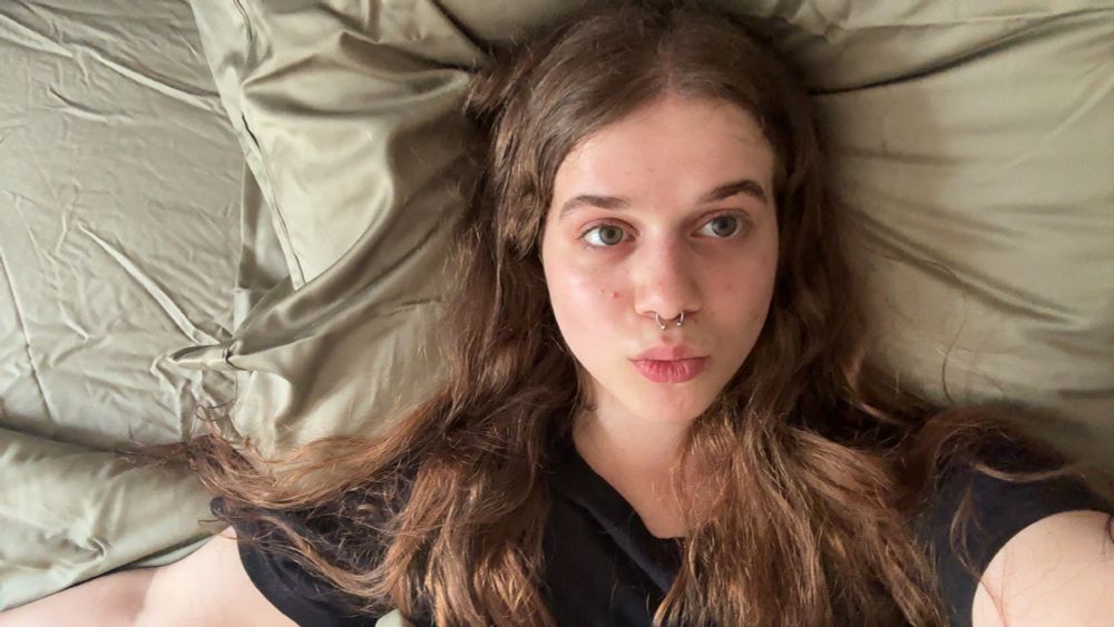 A selfie of me laying on a bed with creased sage green sheets and pillowcase. I'm wearing a black shirt, I have my hair down, and I'm making a duck face.