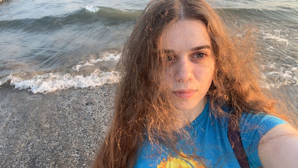 A selfie of me with my hair down in a blue t-shirt standing in front of the lake