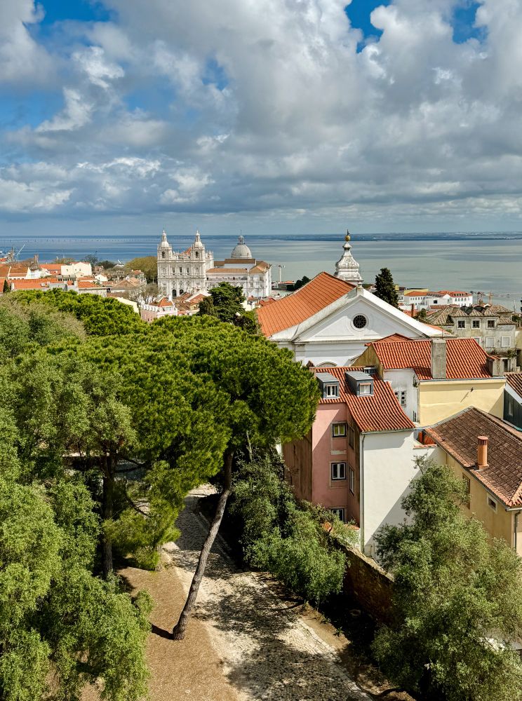 A sunny view looking towards the coast from Lisbon Castle. A cathedral can be seen in the distance, with the Tagus river behind, some rooftops in the middleground, and green trees in the foreground. The sky is blue but fairly cloudy.