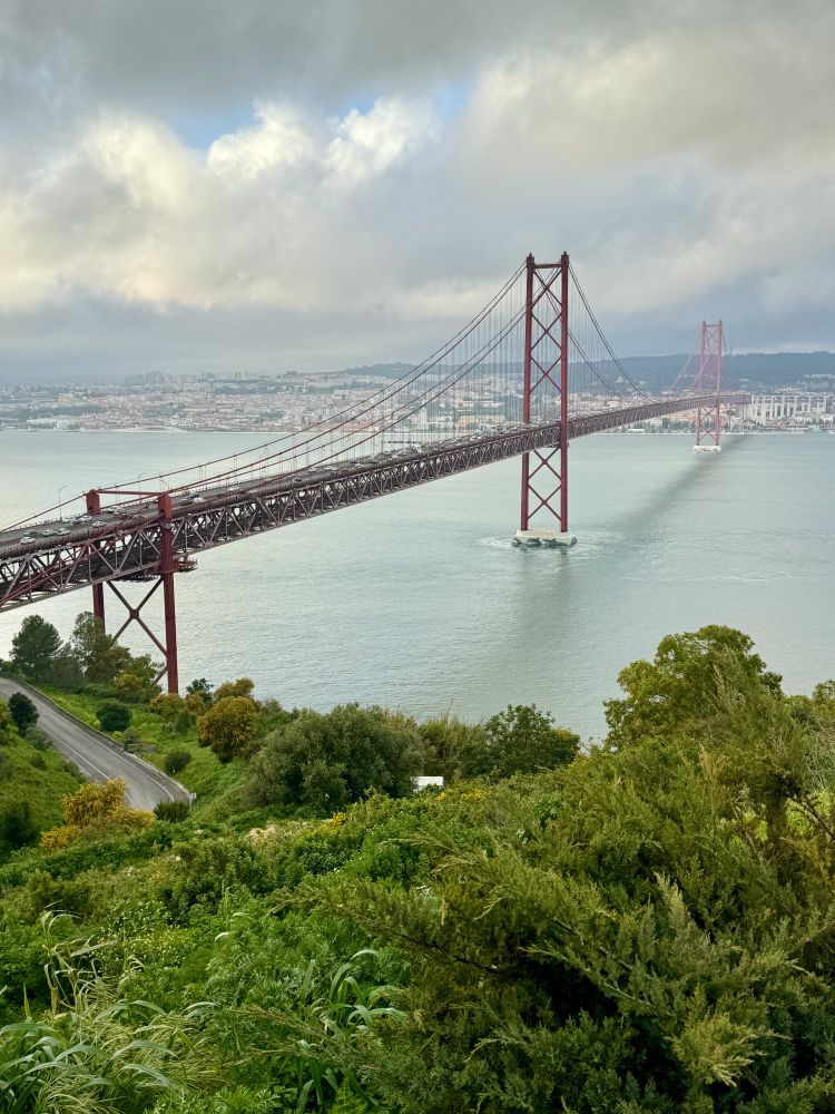 The 25th April bridge, painted the same International Orange as the Golden Gate Bridge, stretches across the Tagus river from the middleground into the background, while foliage and part of a curved road fills the foreground bottom third.