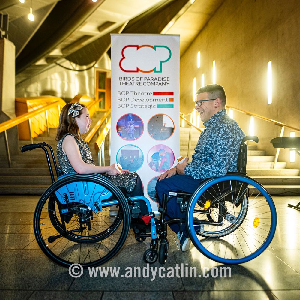 Two smiling people using wheelchairs face each other in front of a banner for Birds of Paradise Theatre Company