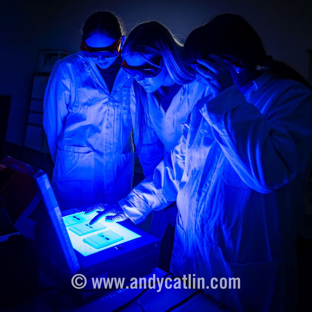 A group of young people in lab coats and protective glasses are gathered around an illuminated light box which is displaying laboratory test results