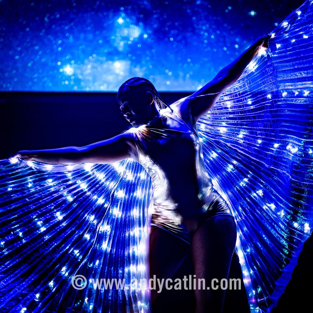 A dancer onstage during a performance with their arms outstretched holding a cloth with embedded lights