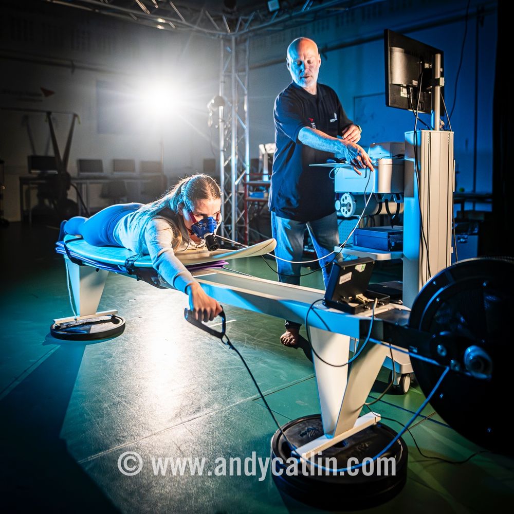 A person lying on a surf board inside a gym is taking part in a fitness check with a researcher standing beside them who is monitoring their progress on a computer.