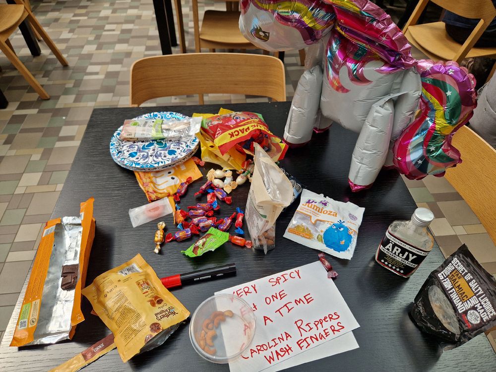 An array of snacks and sweets including one with a written warning poster: "VERY SPICY. EAT ONE A TIME. CAROLINA RIPPERS. WASH FINGERS." Plus two types of Finnish salmiakki which should have a warning, and a big rainbow unicorn balloon. 