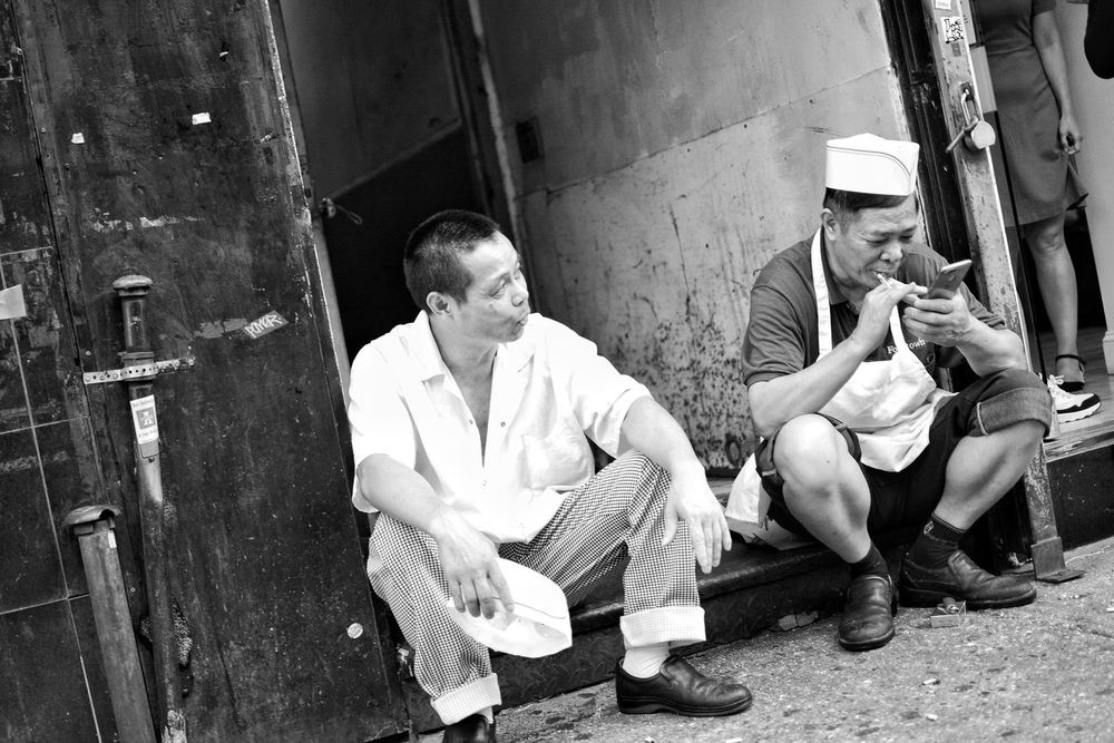 Two men sit outside a shop, one using a smartphone and smoking, the other looking on, Chinatown, Manhattan, New York.