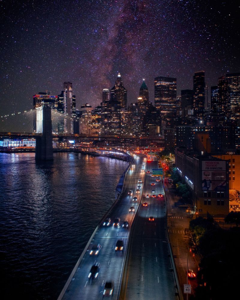 A city skyline at night featuring a brightly lit bridge, moving highway traffic, and a star-filled sky. Featuring the Brooklyn Bridge, FDR Drive, the Manhattan Skyline facing FiDi, composited with a sky view taken in North-South Lake. Two Bridges, Manhattan, New York.
