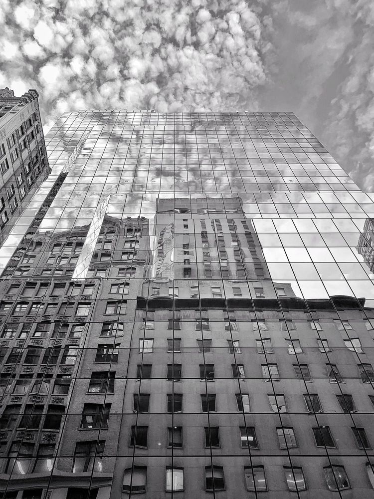A mirrored building in Midtown Manhattan, New York, reflects the surrounding architecture and cloud-filled sky.