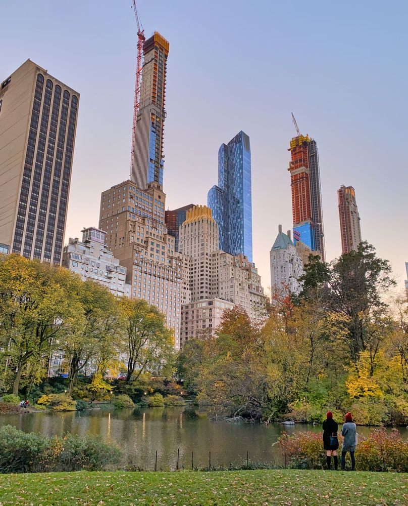Two people stand by a pond surrounded by trees with skyscrapers in the background in Central Park, New York, New York.
