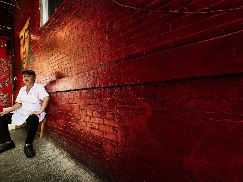 A person sits on a chair beside a red brick wall with a mural of a face on it, Chinatown, Manhattan, New York.